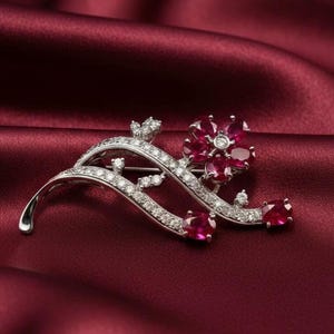 以下が含まれることがあります： 銀色のブローチ、花のモチーフ。赤い宝石の花と透明なクリスタルが特徴です。深い赤色のサテンの背景に配置されています。