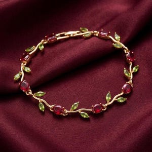May include: Gold-toned bracelet featuring oval red gemstones and small green leaf-shaped accents. The bracelet is set against a deep red fabric background, creating a luxurious aesthetic. The design is reminiscent of a vine.