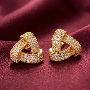 May include: A pair of gold-toned stud earrings shaped like triangular knots. Each earring features a textured surface completely encrusted with small, clear, sparkling stones that cover the entire visible metal frame of the knot design.