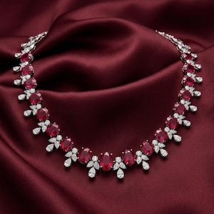 May include: A necklace featuring a series of oval-cut red rubies set in silver-toned metal, accented with small, clear, pear-shaped and marquise-cut white stones arranged in clusters between each ruby, creating a continuous, symmetrical floral pattern.