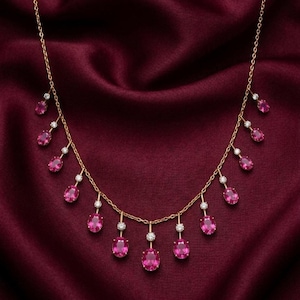 May include: A gold necklace with a delicate chain design. The necklace features multiple pink oval-shaped gemstones and small white gemstones. The necklace is displayed on a deep red satin background.