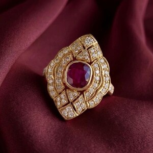 May include: A gold ring featuring a central cushion-cut deep red ruby surrounded by a decorative frame of small, clear pavé-set diamonds. The metal setting has a textured, intricate design that extends outward from the central gemstone.