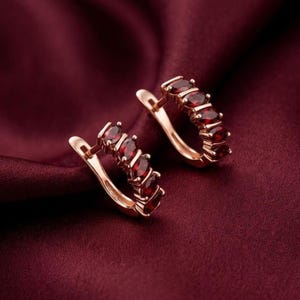 May include: A pair of rose gold hoop earrings with oval-cut, deep red gemstones. The earrings are set against a burgundy fabric background, highlighting the jewelry's color and design. The gemstones are evenly spaced along the front of the hoops.