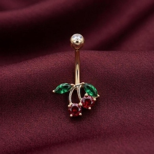 May include: A gold-colored belly button ring featuring a cherry design. The cherries are represented by two red gemstones, with green leaves and a small clear gemstone at the top. The jewelry is set against a burgundy background.