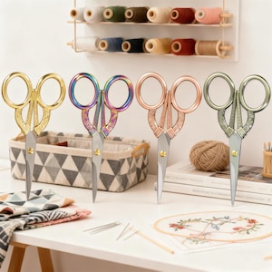 May include: Four decorative embroidery scissors in gold, iridescent, copper, and silver tones. The scissors have ornate handle designs and sharp blades. A basket, spools of thread, and embroidery supplies are also visible.