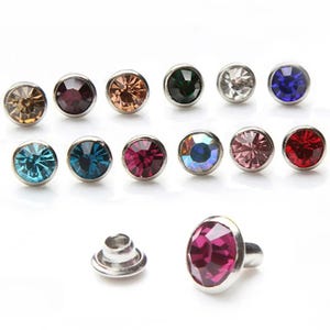 May include: A selection of colourful rhinestone studs in shades like brown, purple, orange, green, clear, and blue. The studs have a silver metal base. One stud is oval-shaped and features a pink rhinestone.