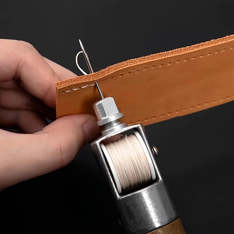 Op de afbeelding: Close-up van een lederen ambachtelijk project in uitvoering. Een handgereedschap met een naald doorboort een bruinleren riem. De tool heeft een zilveren behuizing en een houten handvat, met een spoel wit garen.