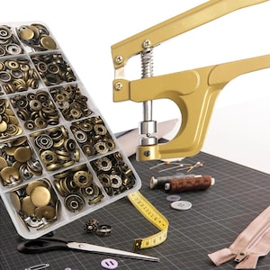 Puede incluir: Un organizador de plástico transparente lleno de varios cierres de presión de color latón antiguo. Una herramienta de prensa de broches de color dorado está colocada a la derecha. También se ven suministros de costura adicionales, como tijeras, una cinta métrica e hilos.