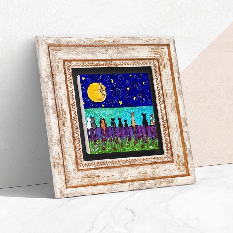 May include: Framed artwork featuring a whimsical scene of cats perched on a fence under a starry night sky with a crescent moon. The artwork has a white wooden frame. The cats are various colors, and the fence is purple.
