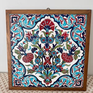 May include: A square decorative tile with a floral pattern in red, blue, and green, set within a wooden frame. The design is set against a white background. The frame provides a warm contrast to the vibrant colours of the artwork.