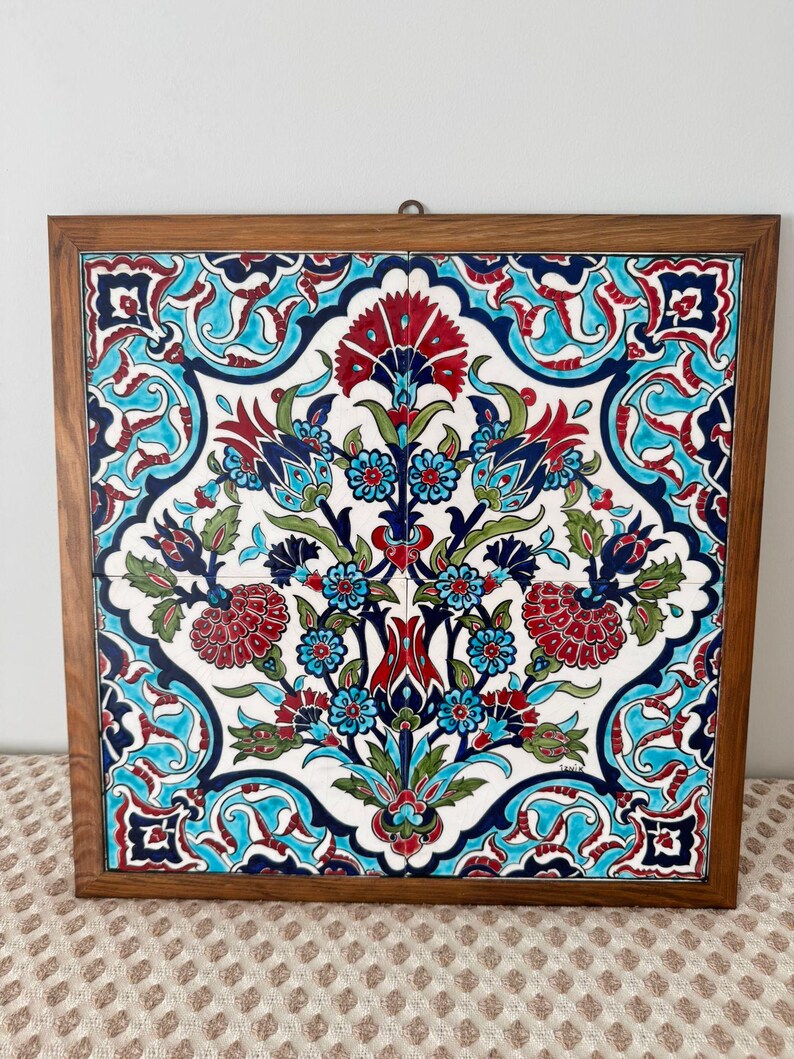 May include: A square decorative tile with a floral pattern in red, blue, and green, set within a wooden frame. The design is set against a white background. The frame provides a warm contrast to the vibrant colours of the artwork.