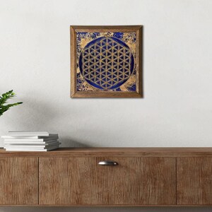 K&ouml;nnte beinhalten: Ein gerahmtes Kunstwerk mit einem goldenen geometrischen Muster, der Blume des Lebens, vor einem tiefblauen Hintergrund. Das Kunstwerk ist in einem Holzrahmen eingefasst und erzeugt einen auff&auml;lligen Kontrast.