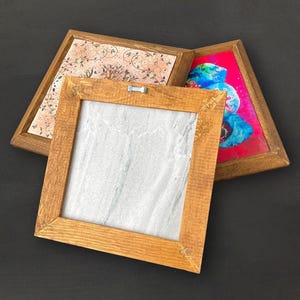 K&ouml;nnte beinhalten: Drei gerahmte Kunstwerke mit Holzrahmen. Eines zeigt ein wei&szlig;es und graues Marmormuster, ein weiteres ein rosa-beiges Blumenmuster und das dritte ein farbenfrohes abstraktes Muster. Die Rahmen sind aus hellbraunem Holz.