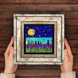 May include: A framed artwork featuring a whimsical night scene. A yellow moon and a sleeping cat are in the night sky. Below, a purple fence is lined with various cat silhouettes. The artwork is in a white distressed frame.