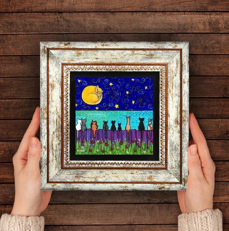 May include: A framed artwork featuring a whimsical night scene. A yellow moon and a sleeping cat are in the night sky. Below, a purple fence is lined with various cat silhouettes. The artwork is in a white distressed frame.