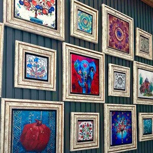 Puede incluir: Una colecci&oacute;n de obras de arte enmarcadas con varios dise&ntilde;os coloridos, incluyendo una granada, un elefante y motivos florales. Los marcos son de madera clara envejecida, y las obras se exhiben en una pared de color verde azulado oscuro.