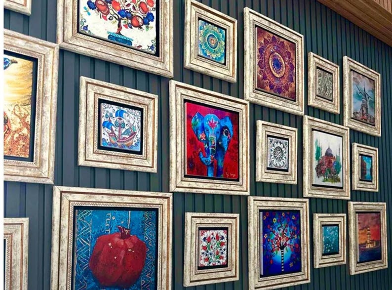 Puede incluir: Una colecci&oacute;n de obras de arte enmarcadas con varios dise&ntilde;os coloridos, incluyendo una granada, un elefante y motivos florales. Los marcos son de madera clara envejecida, y las obras se exhiben en una pared de color verde azulado oscuro.