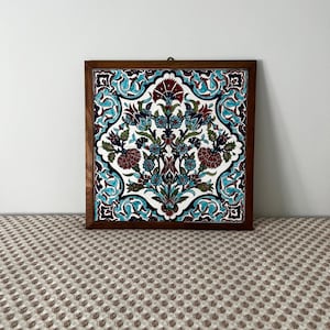 May include: Square decorative tile with a floral design in a wooden frame. The tile features a white background with a symmetrical pattern of teal, burgundy, and blue flowers and leaves. The frame is a dark brown colour.