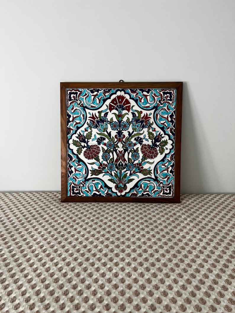 May include: Square decorative tile with a floral design in a wooden frame. The tile features a white background with a symmetrical pattern of teal, burgundy, and blue flowers and leaves. The frame is a dark brown colour.