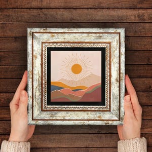 Op de afbeelding: Een ingelijste kunstdruk met een minimalistisch landschap met een zon. Het kunstwerk gebruikt een warm kleurenpalet van oranje, roze en blauw tegen een crèmekleurige achtergrond. De lijst is wit met een verweerde afwerking.
