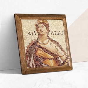 Può includere: Arte a mosaico incorniciato raffigurante una donna con una corona d'alloro e una collana. Il mosaico è composto da piccole tessere colorate, inserite in una cornice di legno. L'opera presenta un testo greco antico. La tavolozza dei colori è calda, con marroni, ori e creme.