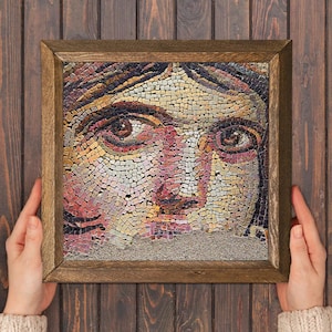 Puede incluir: Una obra de arte en mosaico enmarcada que representa un rostro humano. El mosaico está compuesto por pequeñas baldosas de colores en tonos rosa, amarillo, marrón y morado. El marco es de madera. La obra está sostenida por dos manos.