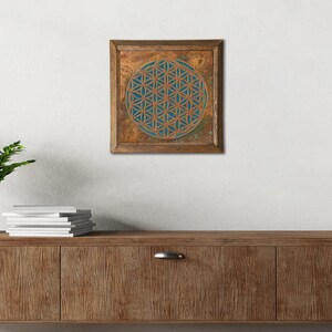 May include: Square wall art featuring a geometric flower of life design. The artwork has a teal and gold color scheme, with a wooden frame. The flower of life is a sacred geometry symbol.
