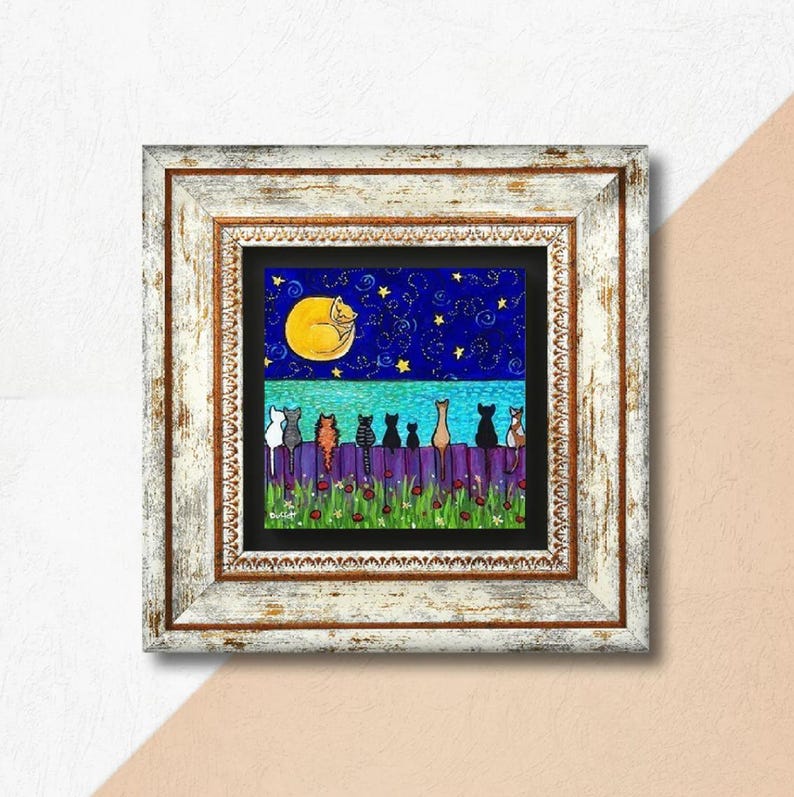 May include: A framed art print depicts a whimsical night scene. A yellow moon with a sleeping cat hangs in a starry blue sky above a purple fence. Several cats of various colors sit on the fence, with red flowers below.