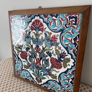 May include: A framed ceramic tile with a floral design. The artwork features a white background with a pattern of red, blue, and teal flowers and leaves. The frame is made of wood.