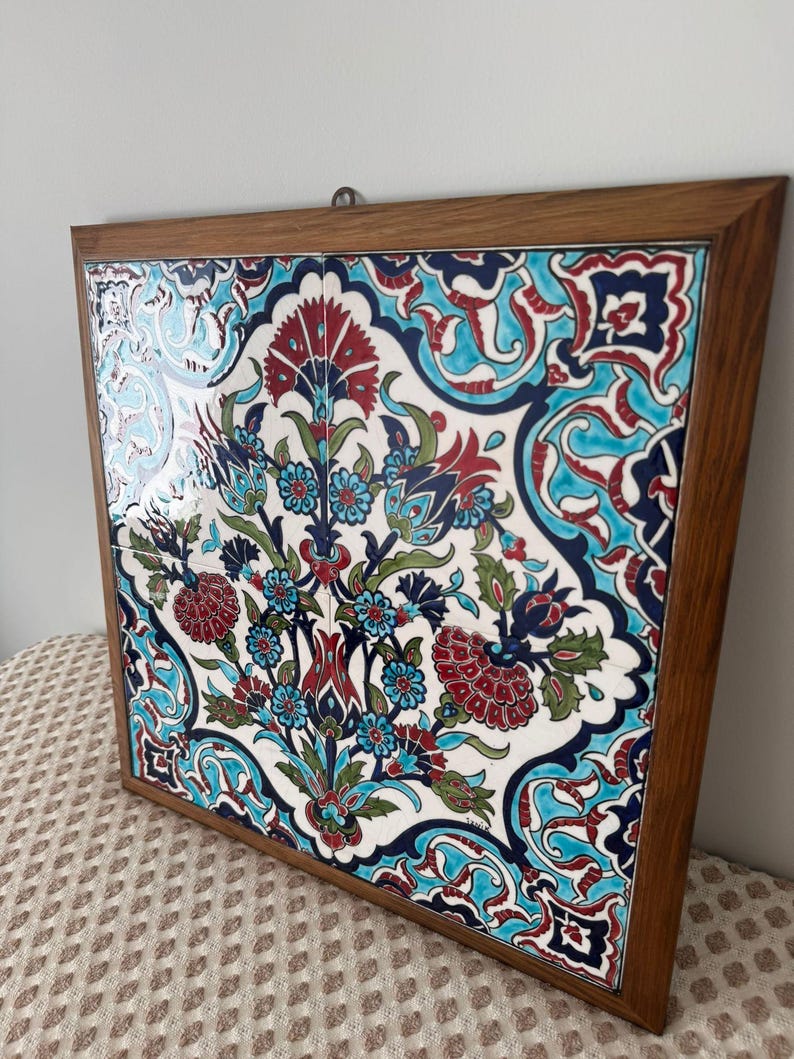 May include: A framed ceramic tile with a floral design. The artwork features a white background with a pattern of red, blue, and teal flowers and leaves. The frame is made of wood.