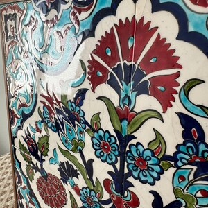 May include: A framed ceramic tile panel with a floral design. The artwork features red, blue, green, and turquoise colours against a white background. The wooden frame surrounds the tiles. The panel is a decorative piece.