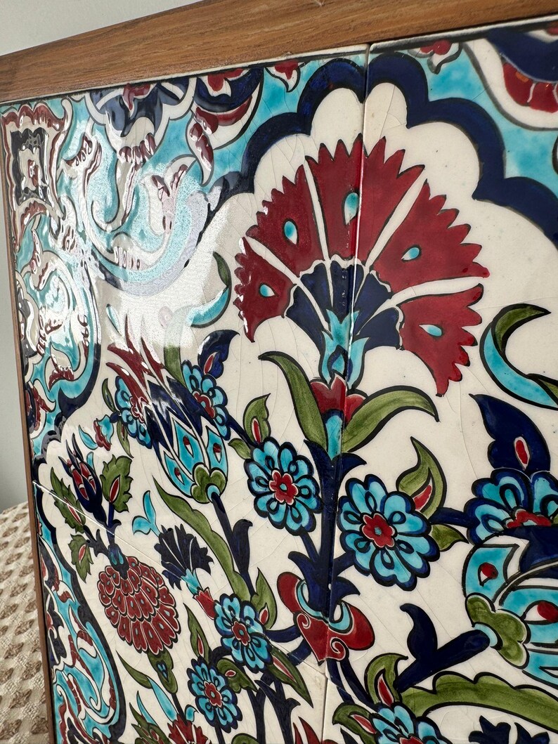 May include: A framed ceramic tile panel with a floral design. The artwork features red, blue, green, and turquoise colours against a white background. The wooden frame surrounds the tiles. The panel is a decorative piece.
