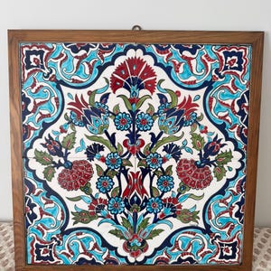 May include: Square ceramic tile with a floral design in a wooden frame. The tile features a white background with a symmetrical pattern of red, blue, and green flowers and leaves. The frame is a warm brown colour.