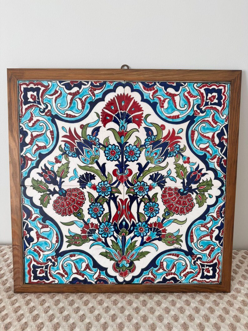 May include: Square ceramic tile with a floral design in a wooden frame. The tile features a white background with a symmetrical pattern of red, blue, and green flowers and leaves. The frame is a warm brown colour.
