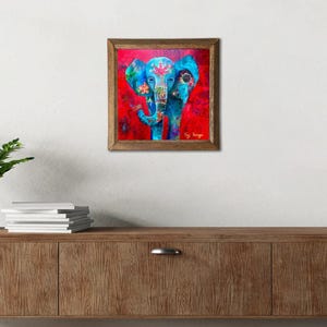 Puede incluir: Una impresi&oacute;n art&iacute;stica cuadrada enmarcada que presenta un elefante azul con patrones florales y geom&eacute;tricos sobre un fondo rojo. El elefante tiene orejas grandes y una trompa. La obra est&aacute; firmada en la esquina inferior derecha.