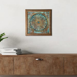 K&ouml;nnte beinhalten: Quadratische Wandkunst mit Holzrahmen und einem floralen Design in T&uuml;rkis und Gold. Das Kunstwerk zeigt ein kreisf&ouml;rmiges Muster mit detaillierter, strukturierter Optik. Der Rahmen hat eine warme braune Farbe und das St&uuml;ck ist an einer wei&szlig;en Wand befestigt.