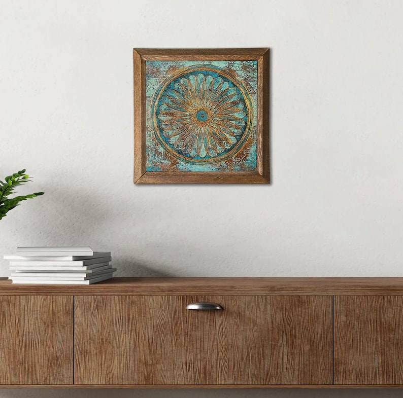 K&ouml;nnte beinhalten: Quadratische Wandkunst mit Holzrahmen und einem floralen Design in T&uuml;rkis und Gold. Das Kunstwerk zeigt ein kreisf&ouml;rmiges Muster mit detaillierter, strukturierter Optik. Der Rahmen hat eine warme braune Farbe und das St&uuml;ck ist an einer wei&szlig;en Wand befestigt.
