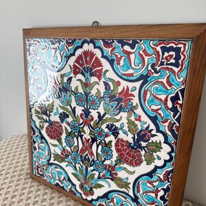 May include: A framed decorative tile with a floral pattern in red, blue, and green on a white base. The square tile is encased in a wooden frame and has a metal hook for hanging. The design features stylized flowers and foliage.