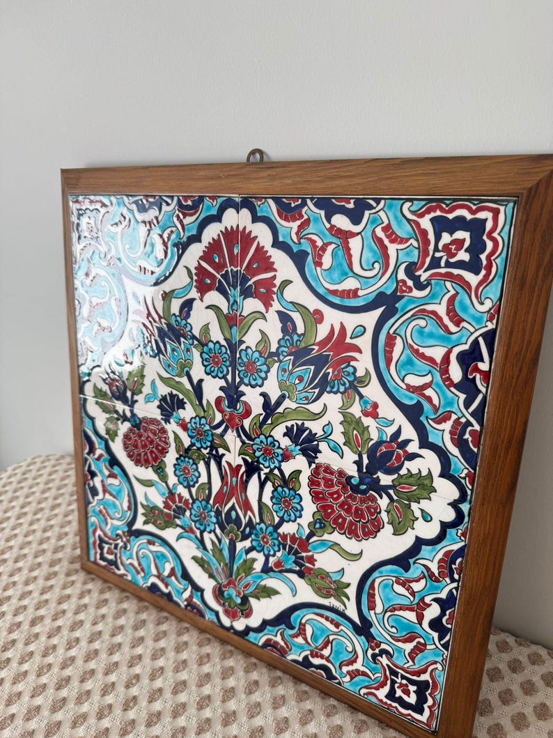 May include: A framed decorative tile with a floral pattern in red, blue, and green on a white base. The square tile is encased in a wooden frame and has a metal hook for hanging. The design features stylized flowers and foliage.