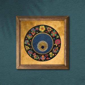 May include: A framed artwork with a circular design. It features a blue eye motif at the centre, encircled by vibrant floral patterns on a black base. The artwork is set against a gold-toned background within a wooden frame, ideal for home decor.