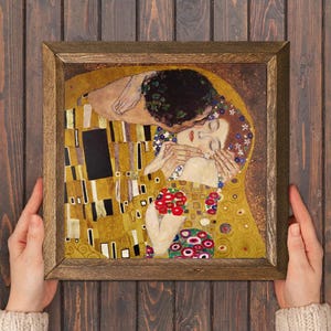 May include: A framed print of Gustav Klimt's "The Kiss" depicts a couple embracing, with gold and yellow tones prominent. The artwork is held by hands against a dark wood backdrop, highlighting the intricate details of the painting.