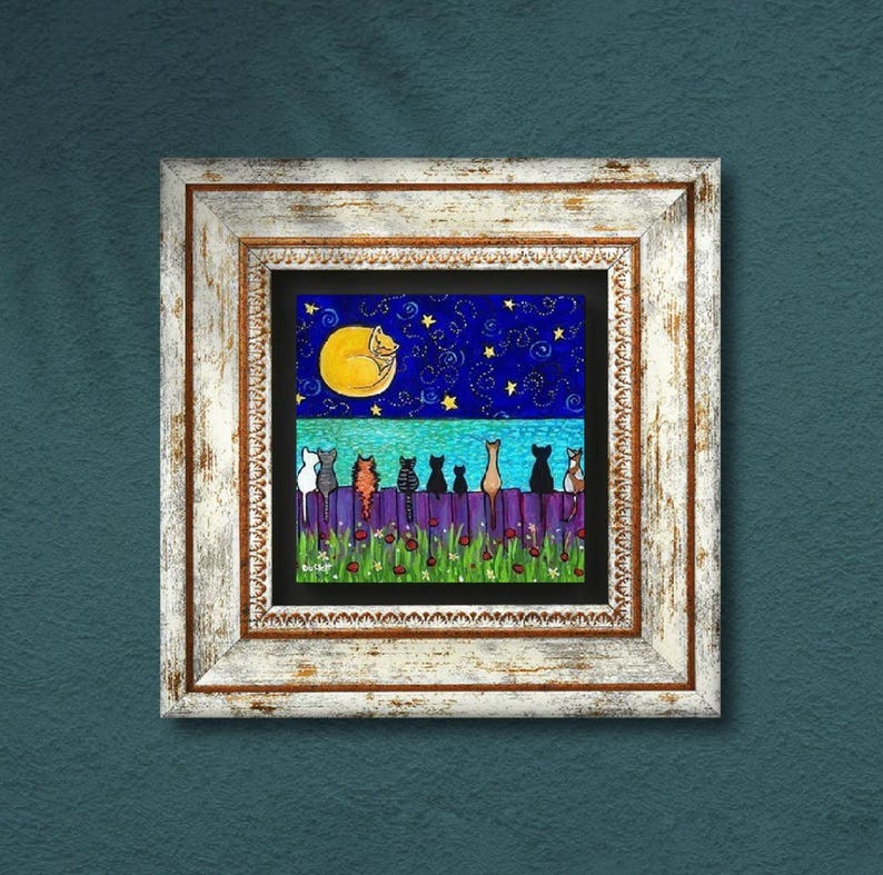May include: A framed artwork depicts a whimsical night scene. A row of stylized cats sits on a purple fence, beneath a starry blue sky and a large yellow moon with a sleeping cat. The frame is white with a distressed finish.