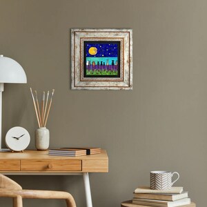 May include: A framed art print featuring a whimsical night scene. The artwork depicts silhouetted cats on a fence under a starry sky and a full moon. The frame is white with a distressed finish, adding a rustic touch to the artwork.