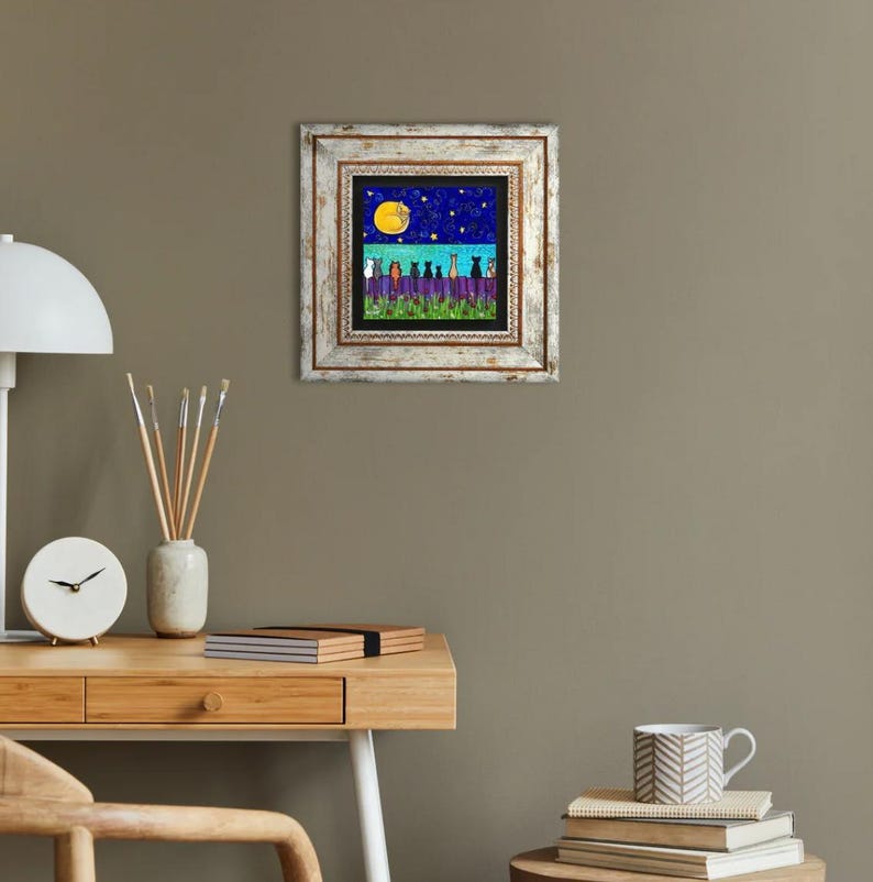 May include: A framed art print featuring a whimsical night scene. The artwork depicts silhouetted cats on a fence under a starry sky and a full moon. The frame is white with a distressed finish, adding a rustic touch to the artwork.