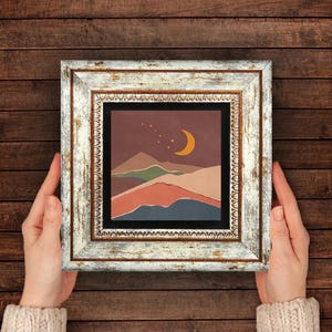 Op de afbeelding: Een ingelijste kunstdruk met een minimalistische landschapsscène. Het kunstwerk toont bergen in bruin-, groen- en blauwtinten onder een nachthemel met een halve maan en sterren. De lijst is wit met een antieke afwerking.