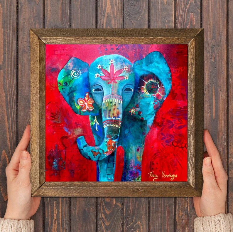 Puede incluir: Una obra de arte enmarcada que presenta un elefante vibrante y estilizado sobre un fondo rojo. El elefante est&aacute; pintado en tonos azules, adornado con motivos florales y de hojas. La obra est&aacute; firmada por Tracy Verdugo.