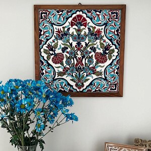 May include: A framed, square ceramic tile with a floral design in red, teal, blue, and green against a white background. The tile is set in a dark brown wooden frame. The artwork is hanging on a white wall.