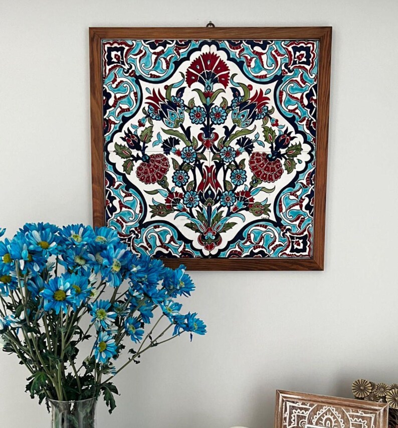 May include: A framed, square ceramic tile with a floral design in red, teal, blue, and green against a white background. The tile is set in a dark brown wooden frame. The artwork is hanging on a white wall.