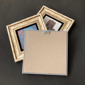 May include: Two framed art pieces with ornate, off-white frames. One features a blue and red design, the other a hand symbol. The back of a third frame is visible, showing a cardboard backing and a metal hanger, all set against a dark background.