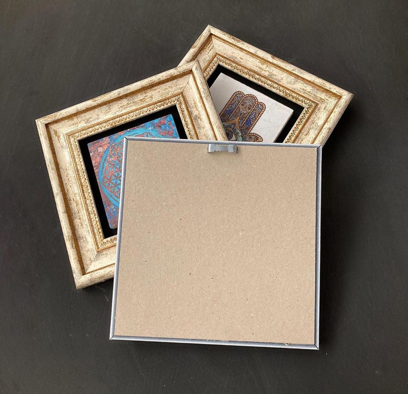 May include: Two framed art pieces with ornate, off-white frames. One features a blue and red design, the other a hand symbol. The back of a third frame is visible, showing a cardboard backing and a metal hanger, all set against a dark background.